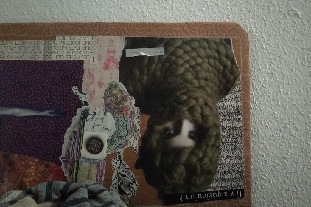Une photo d'un collage sur carton renversé, contre un mur en crépi blanc. Sur le collage, une figure à l'envers dans une cagoule de grosse laine verdâtre et les yeux très maquillés.